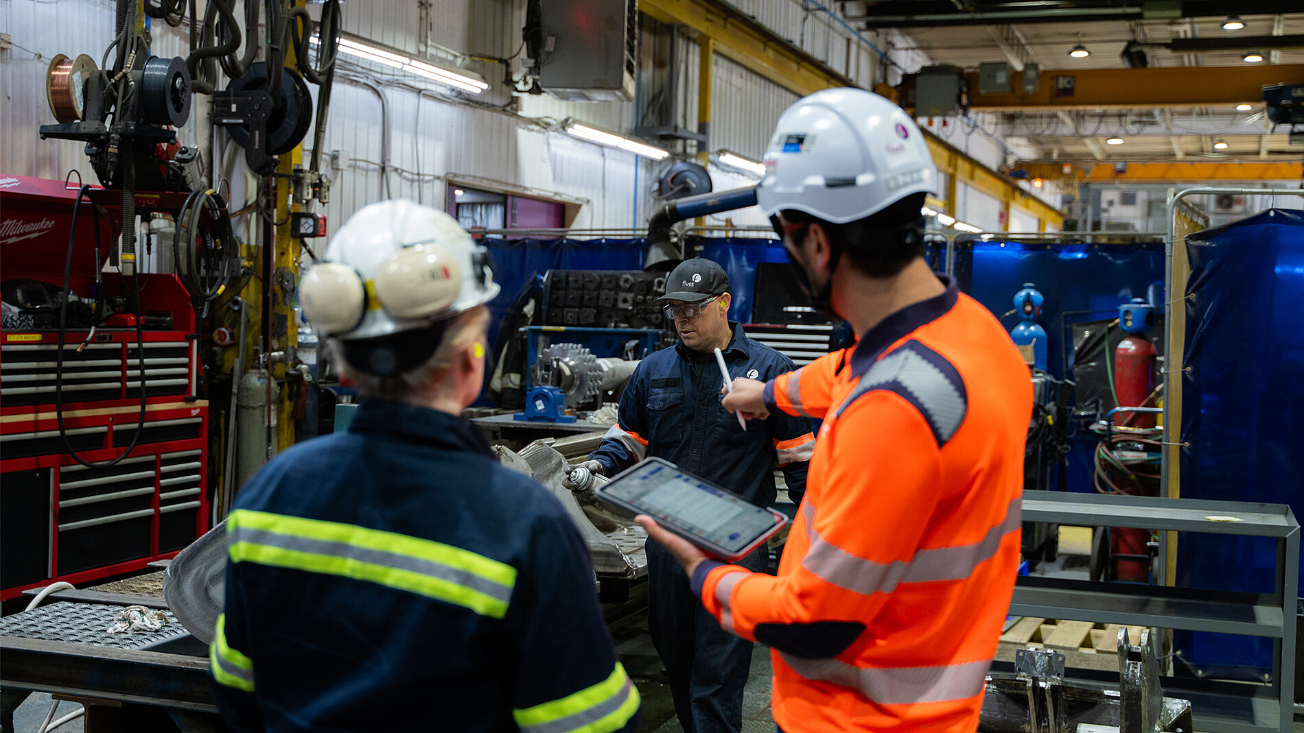 Two employees wearing safety gear review information on a tablet inside an industrial workshop, surrounded by machinery, tools, and manufacturing equipment.